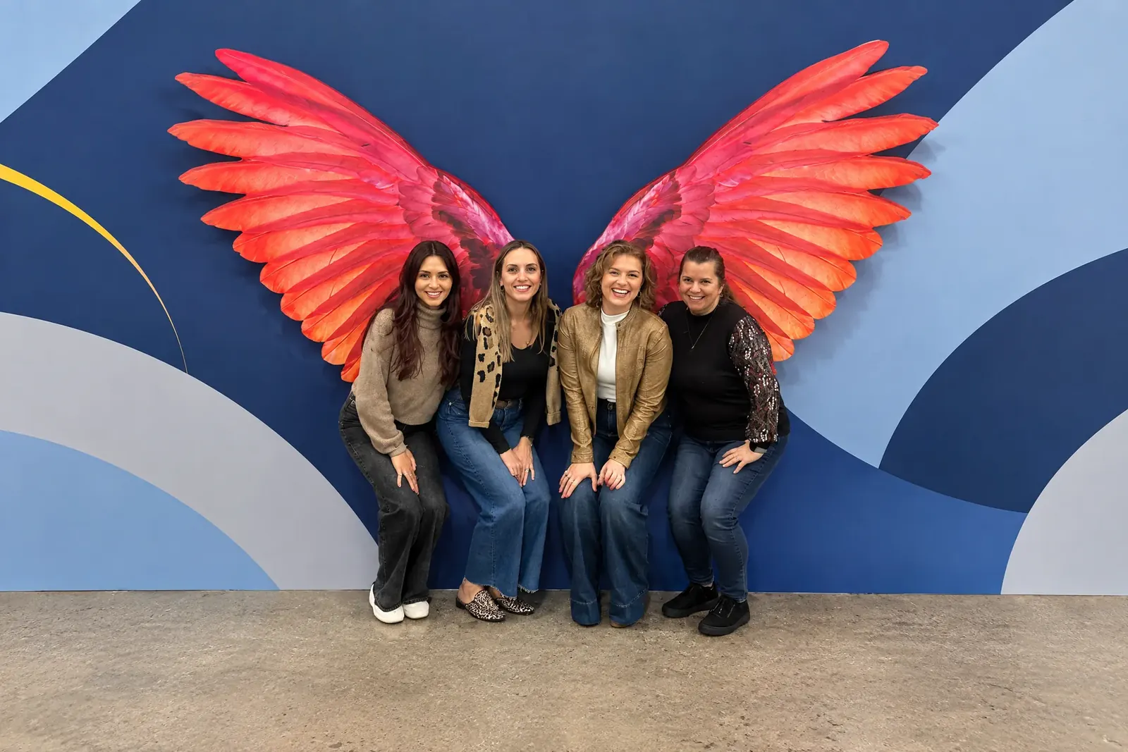 Four Tomegan team members smiling together in front of a colorful wing mural